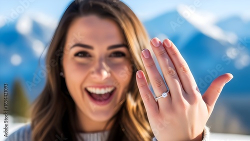 Excited young woman showing off a sparkling new diamond engagement ring with a joyful smile against a scenic snowy mountain backdrop after a romantic proposal