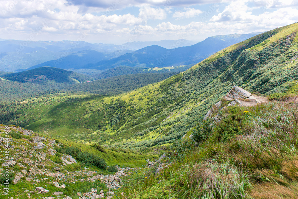 Fototapeta premium Mountain valley landscape with green hills and distant blue peaks under cloudy summer sky