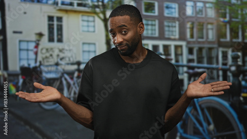 Φωτογραφία Man in black t shirt shrugging with hands raised on a city street near bicycles and canal buildings, bare forearms visible; uncertainty