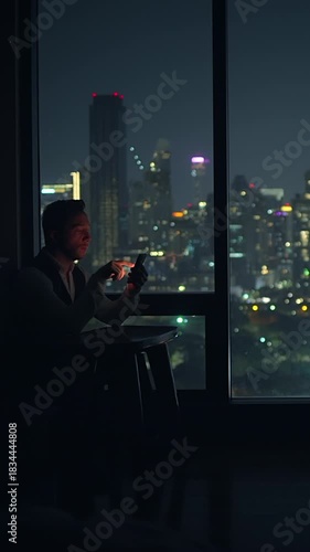 solitary man studies his phone screen with city lights reflecting on hotel window at night