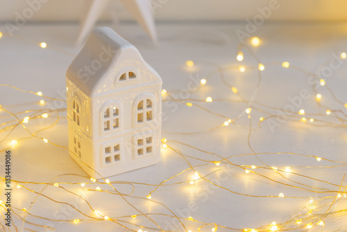 Soft Christmas scene with white ceramic house, glowing warm fairy lights and neutral winter decor. Cozy minimalist holiday background perfect for seasonal designs.
