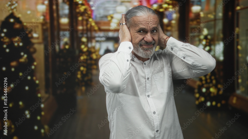Fototapeta premium Man with gray hair and beard covers ears in decorated shopping building corridor holding hands to head in pain; holiday discomfort.
