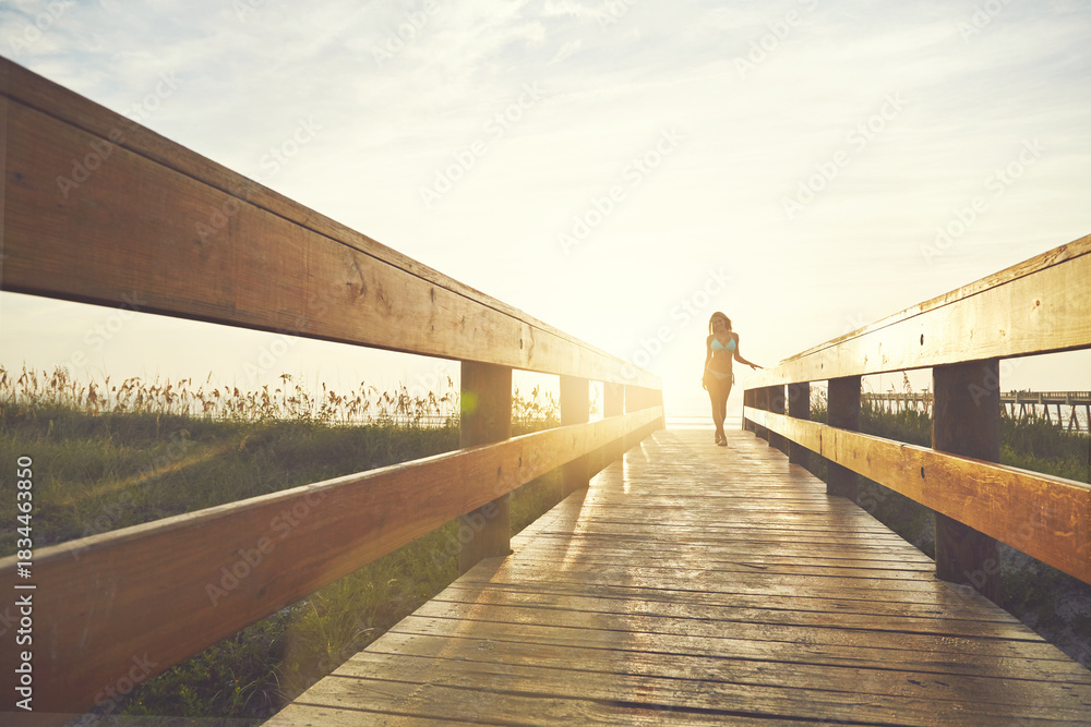 Fototapeta premium Woman, boardwalk and walking at beach with sunshine, below space and vacation for summer travel. Adventure, portrait or person in bikini for coastal holiday, sunrise sky and relax by wood pathway