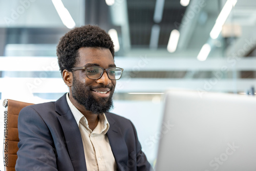 Canvas Print African american businessman smiling at laptop in a modern, sunlit office, focus