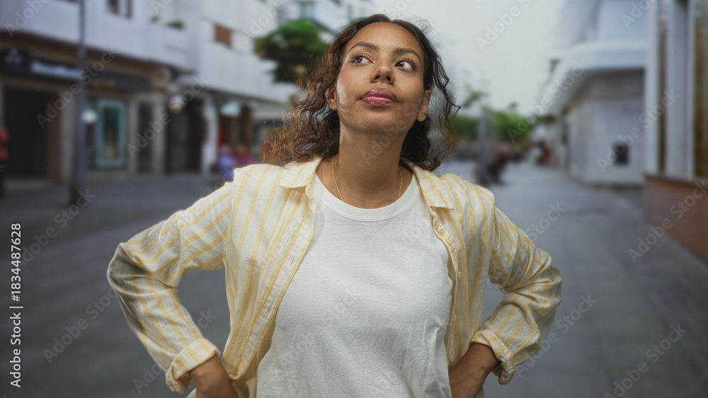 Fototapeta premium Woman touching chin with finger on a city street in a casual striped shirt, glancing upward; thoughtful curiosity.