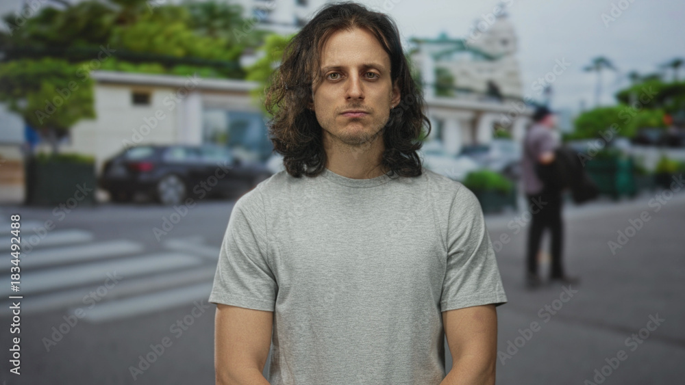 Fototapeta premium Man with long hair standing with arms relaxed on street near crosswalk and parked car; introspection solitude.