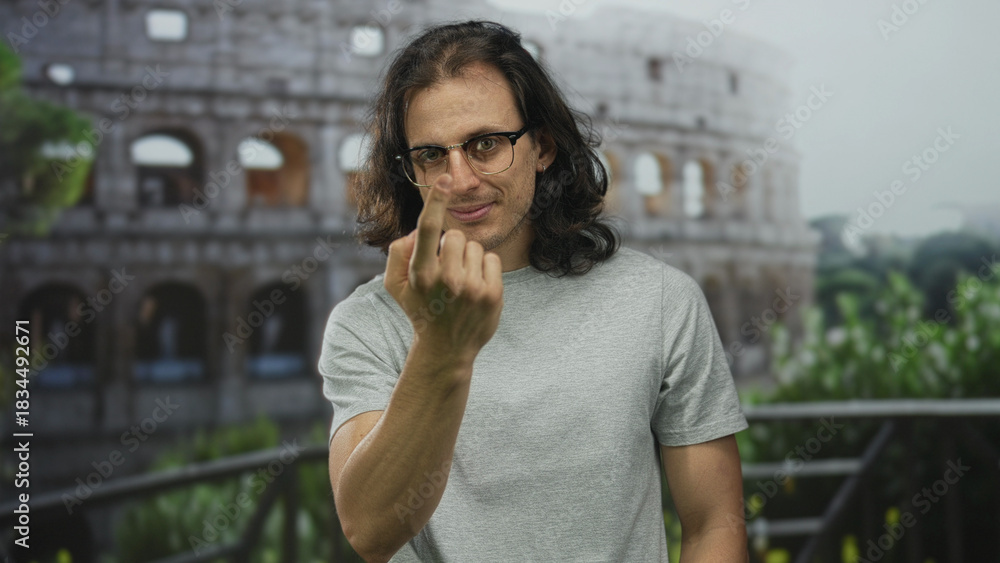 Fototapeta premium Man showing middle finger to camera on a building terrace near the colosseum, tshirt and glasses; defiance provocation insult.