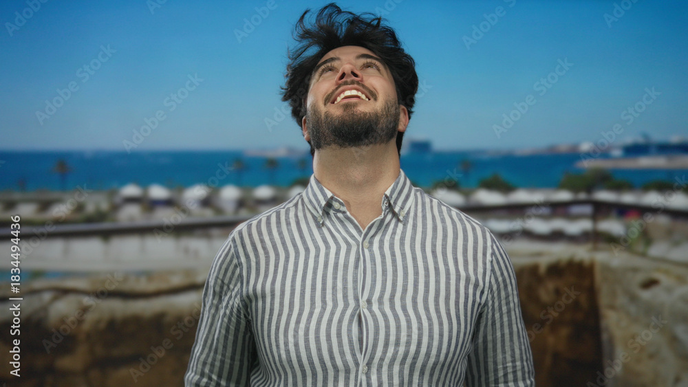 Fototapeta premium Young hispanic man with beard smiling at port with ships in background enjoying a sunny day while wearing a striped shirt and feeling the sea breeze on his face