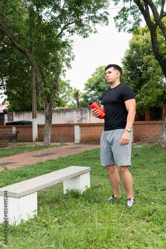Standing man holding a red water bottle at the park.