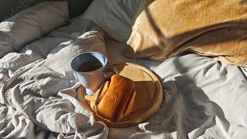 A cup of hot tea with a fresh bun rests on a wooden board placed on soft bedding in warm sunlight. The scene conveys cozy relaxation and a slow, peaceful morning atmosphere
