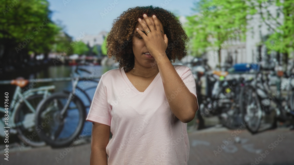 Fototapeta premium Woman holding forehead with hand near temple on busy amsterdam street beside parked bicycles and canal bridge; frustration headache.