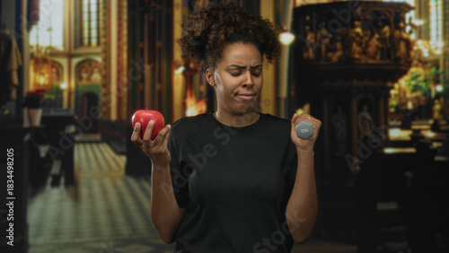 Woman holds red apple and 1kg dumbbell in her hands inside a church building, grimacing; temptation and doubt.