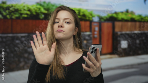 Woman raising hand to stop while glancing down at smartphone at street entrance on sidewalk during daytime; defiance.