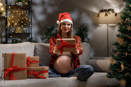 A pregnant woman wearing a Santa hat offers a wrapped gift, celebrating the holidays