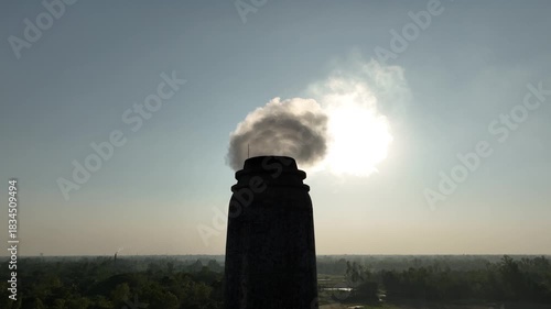 Thick smoke bursts from a brickfield chimney under the blazing sun, filling the sky and quietly harming nearby habitats, showing the harsh impact of industrial pollution.