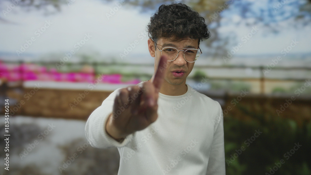 Fototapeta premium Young hispanic man in white sweater and round eyeglasses pointing finger at camera on busy street; skepticism.