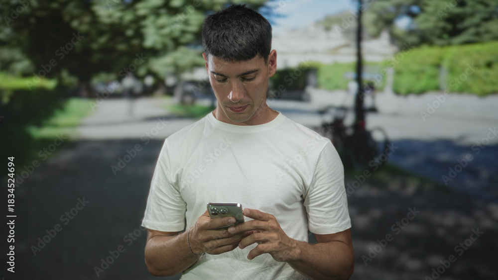 Fototapeta premium Man in white tshirt holding smartphone with hands texting in sunny green park; serene concentration.
