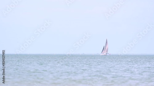A lone sailboat with white sails floats on a calm sea beneath a pale sky.