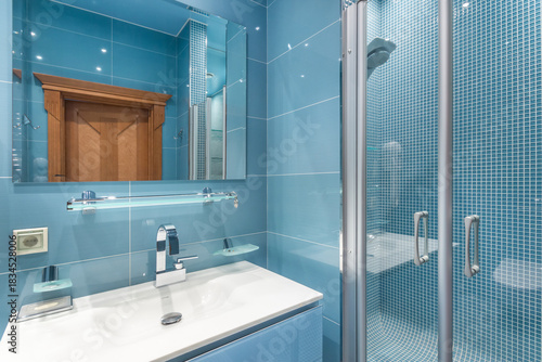 A blue tiled bathroom features a sink with a mirror above, a glass shower, and wooden accents. The design is clean and well-lit, suitable for daily use