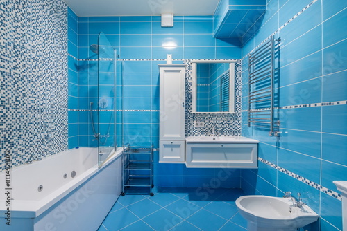 A bathroom with blue tiles features a tub and a separate shower area. A mirror hangs above the sink next to a storage unit. The space is bright and organized