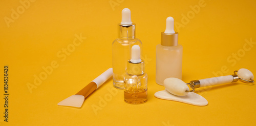 Glass bottles with serums and pipettes stand next to a massage roller and a jade gua sha scraper, a brush for applying a mask