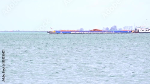 A tugboat and a long barge loaded with orange mounds are visible on calm open water near the horizon.