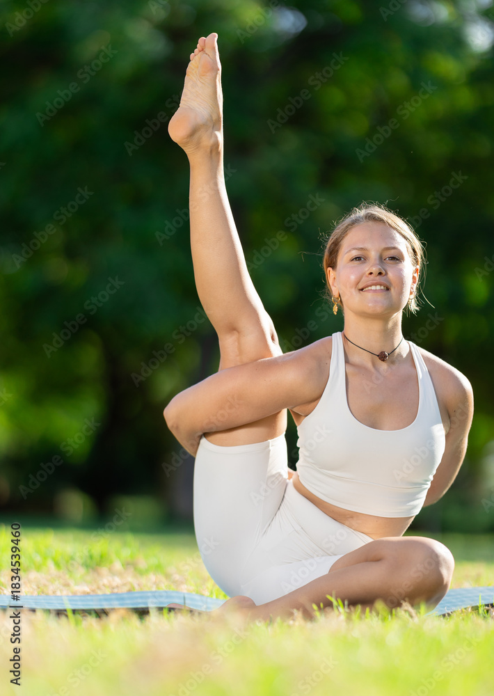 Fototapeta premium Smiling sporty young girl maintaining active lifestyle training in summer sun on bright green meadow, performing dynamic asanas to improve body flexibility, balance and strengthen core..