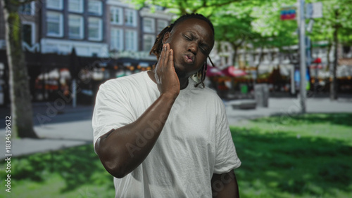 Foto Man with hand on cheek for toothache on street near shops and trees wearing white t shirt; pain seeking help