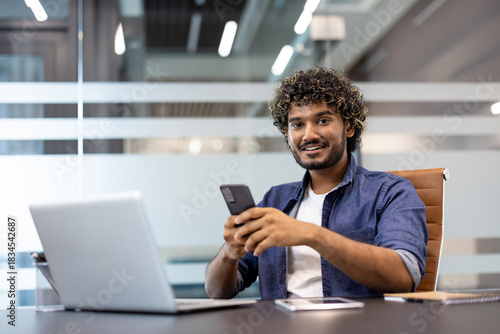 Portrait of a smiling young Indian man sitting at an office desk, holding a phone in his hand and smiling at the camera