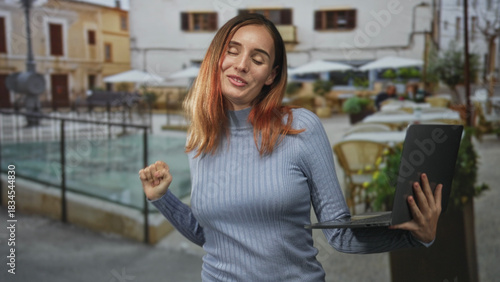 Free Download Woman holding laptop with left hand and fist pumping with right hand on street cafe, smiling and dancing while looking at screen; joy celebration. - GFXLoad