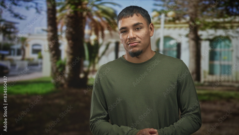 Fototapeta premium Man with hands clasped and a wry grimace, wearing an olive long sleeve shirt, standing near palm trees in front of a sunlit building; skepticism.