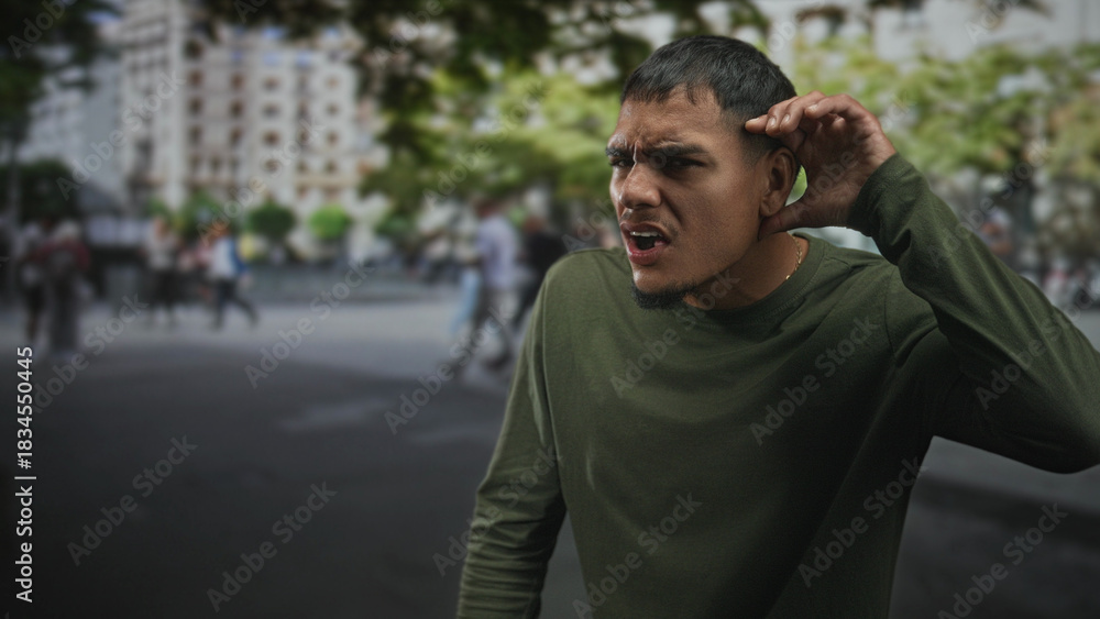 Fototapeta premium Man with visible face and right hand lifted near ear standing center frame among blurred pedestrians and buildings on street; pensive solitude.