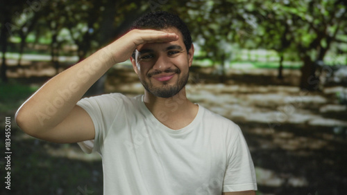Young man wearing white tshirt shades eyes with hand overhead while smiling in green forest sunlight; curiosity optimism adventure.