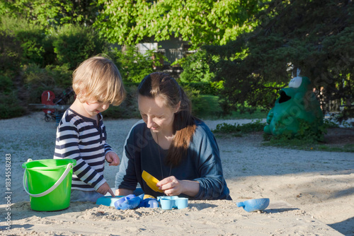 Mother and cute little child boy having fun playing with sand and colorful toys in the park, beautiful summer sunny day in children playground. Selective focus