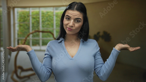 Young hispanic woman stands in an indoor living room gesturing with an expression of indifference and confusion in a relaxed home setting.