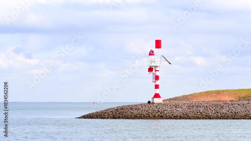 A red and white navigational lighthouse stands on a rocky breakwater under a cloudy sky.