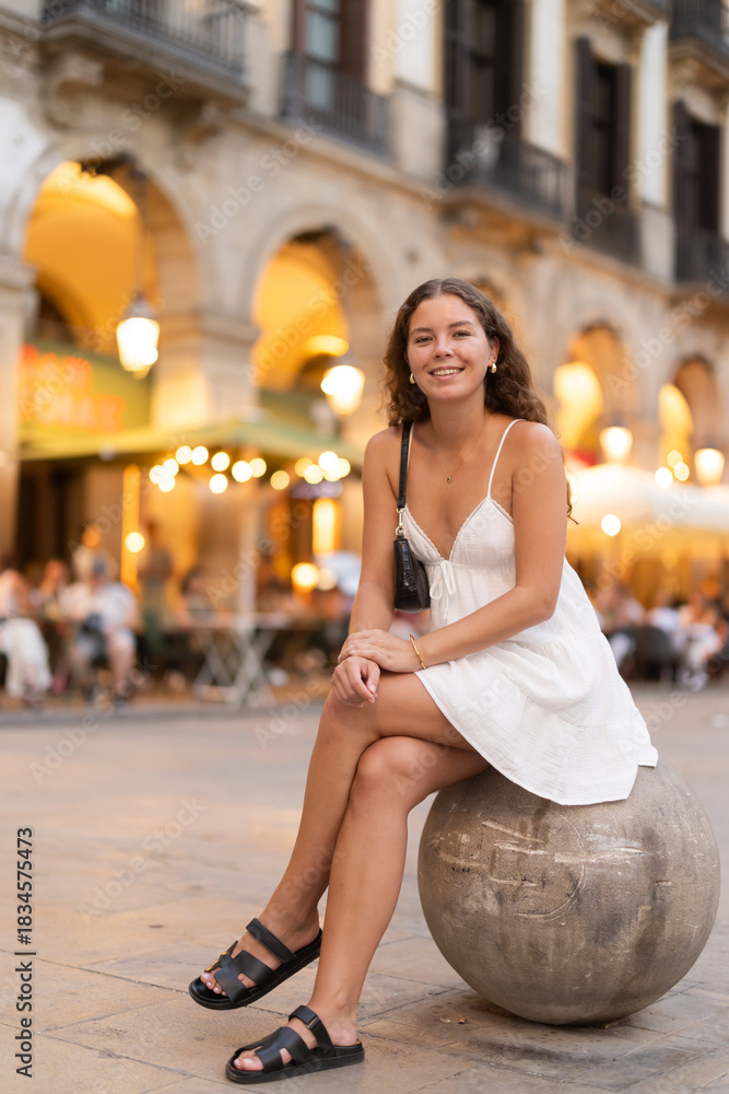 Obraz premium Evening Royal square in Barcelona, girl in sundress is sitting on stone ball, resting on concrete fencing. Electric street lighting, twilight in southern city, evening life in big city