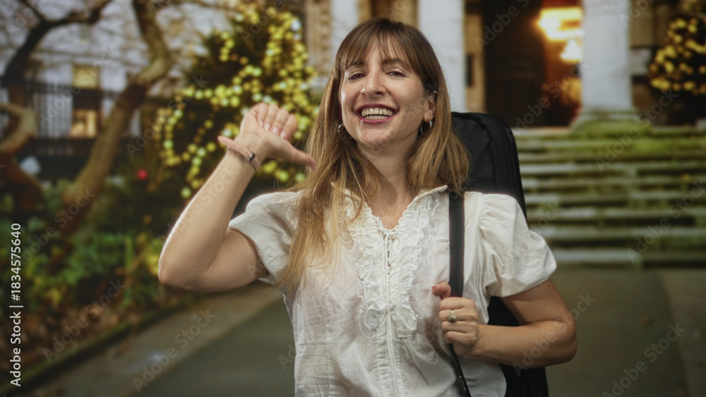 Fototapeta premium Woman points thumb to chest while carrying a guitar case on a city street near building steps; confidence celebration.