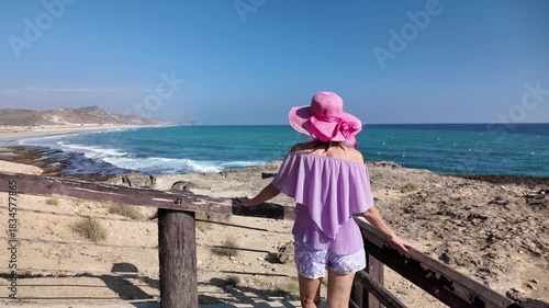 Young girl wearing a pink hat stands at a viewpoint, observing the expansive turquoise ocean and long sandy Mughsayl Beach coastline with mountains under clear blue sky close to Salalah, Dhofar, Oman