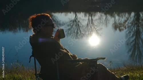 Woman relaxing with hot drink by lake at sunset. Silhouette of a woman sitting in a folding chair on the lake shore, drinking a hot beverage from a mug while enjoying the sunset reflection on water