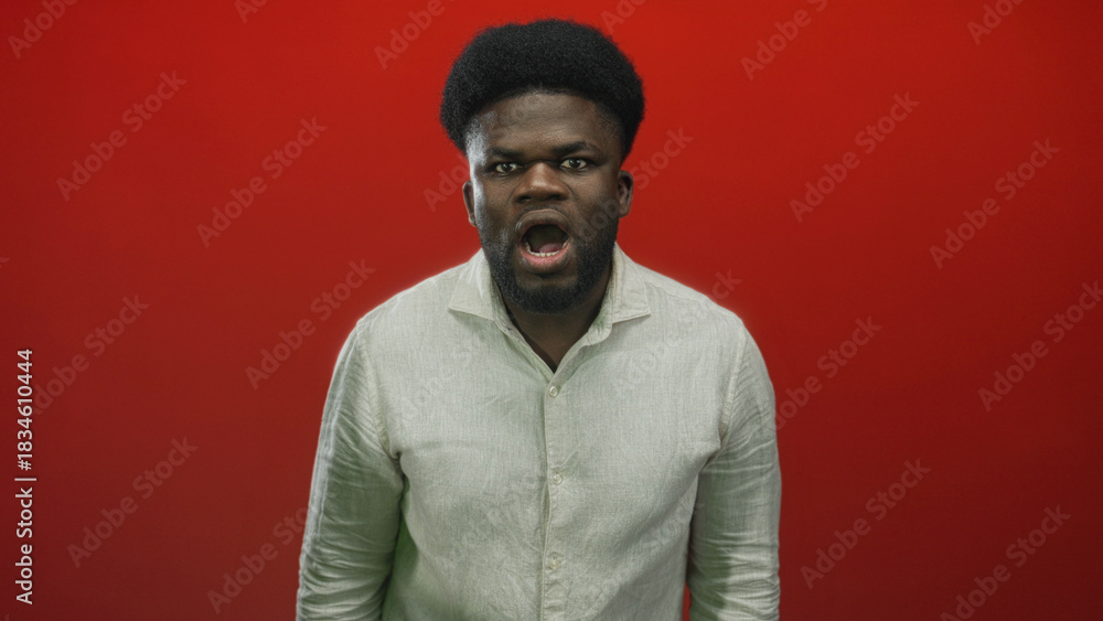 Obraz premium Young african american man shouting with open mouth and intense gaze wearing light linen shirt in studio; anger defiance protest.