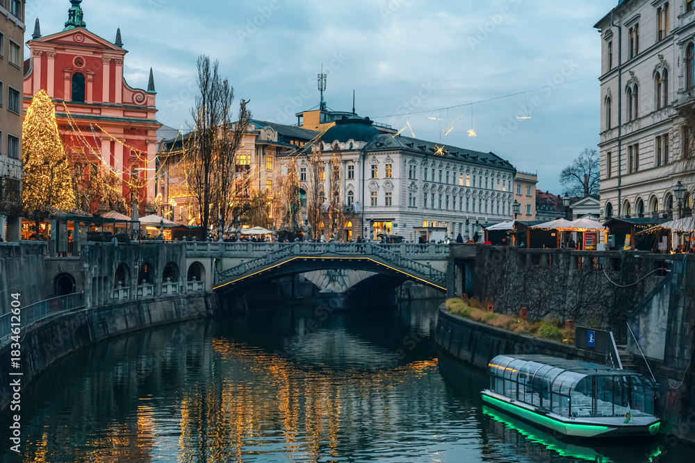 Obraz premium Festive city river with illuminated buildings, decorated trees and glowing reflections at dusk. Ljubljana, Slovenia