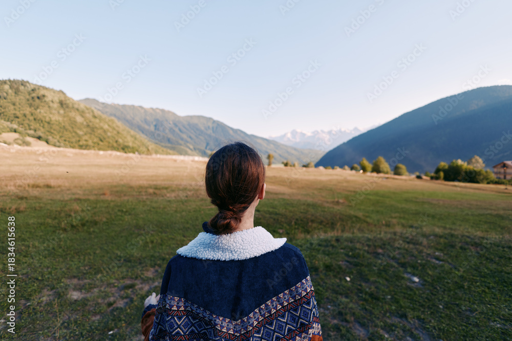 Fototapeta premium Woman back landscape mountains meadow sweater viewed from behind in peaceful nature scene, travel and solitude across a wide valley horizon with scenic outdoors and warm sunlight.