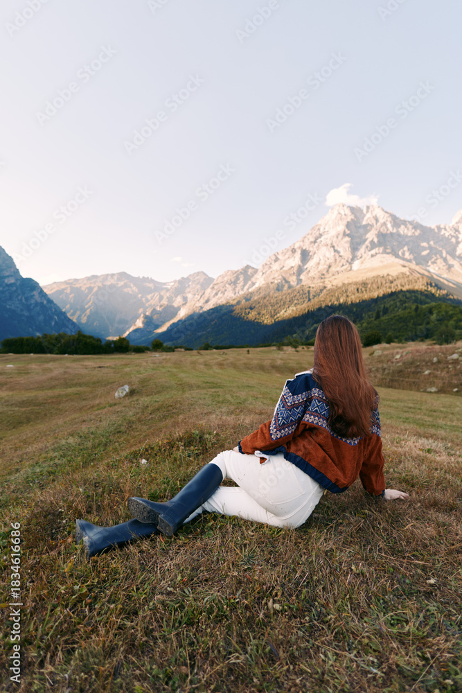 Fototapeta premium Woman sitting in a meadow gazing at majestic mountains, nature landscape during autumn travel and solitude, cozy sweater and boots on grassy valley with serene outdoor scenery and calm.