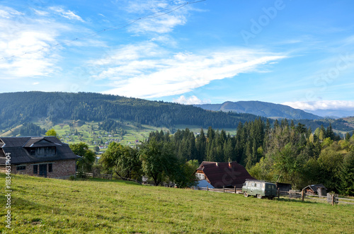 Serene rural landscape featuring a small village nestled in rolling hills, wooden houses, a vintage van, and green meadows under a bright blue sky. Carpathian Mountains, Ukraine