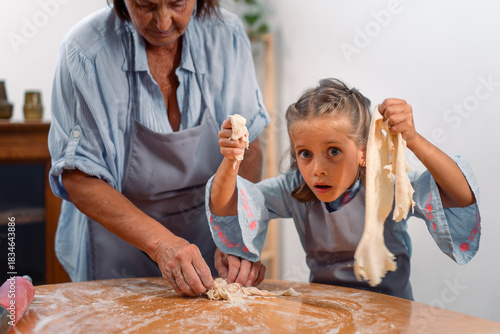 Young girl looking surprised while holding torn dough while baking with grandmother at home kitchen both in aprons having fun and learning together
