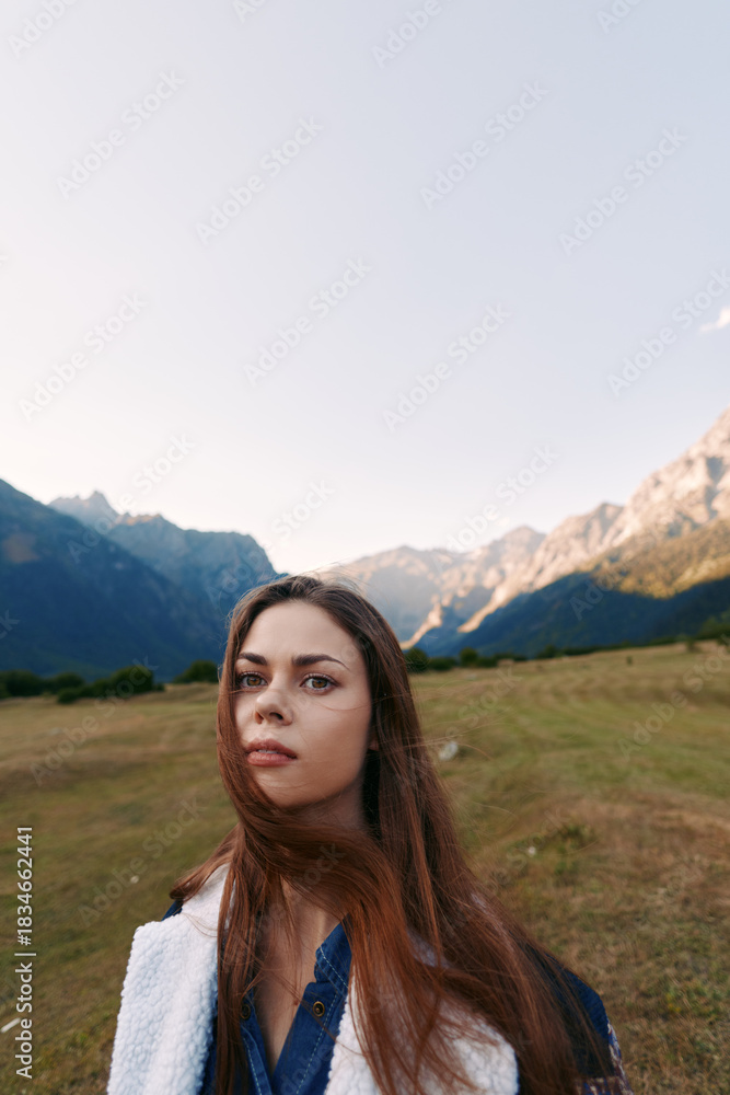 Fototapeta premium woman portrait in mountains meadow, nature landscape outdoors scene with young traveler looking into camera, long hair and thoughtful expression in wide alpine field at golden hour