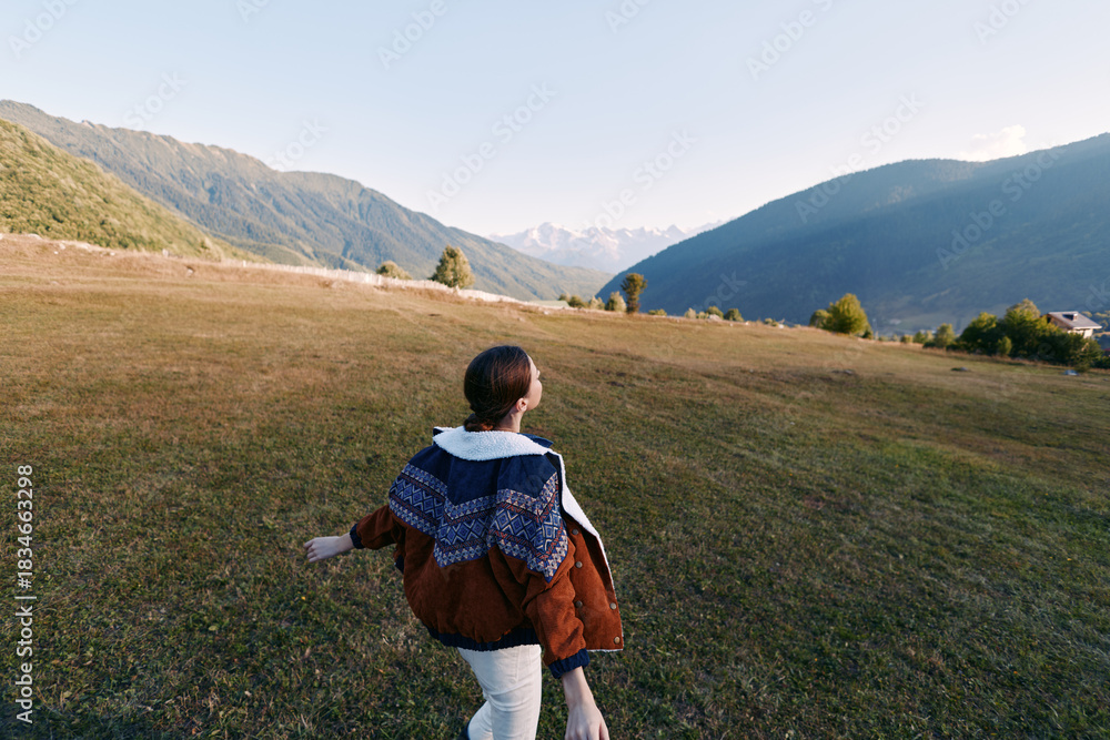 Fototapeta premium Woman walking in a meadow toward mountains across open landscape, nature hiking outdoors travel scene with jacket and shawl, back view capturing freedom and scenic valley peace.