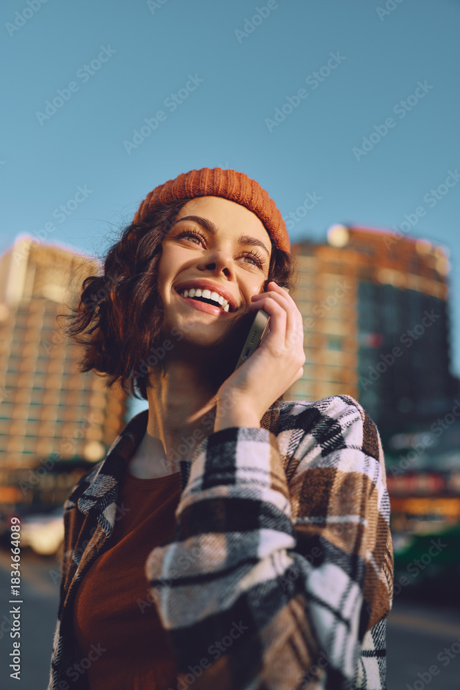 Fototapeta premium Woman on phone smiling in city with beanie and flannel, earbuds in sunlight. Candid lifestyle portrait at golden hour glow, authenticity and mindful living with emotional storytelling.