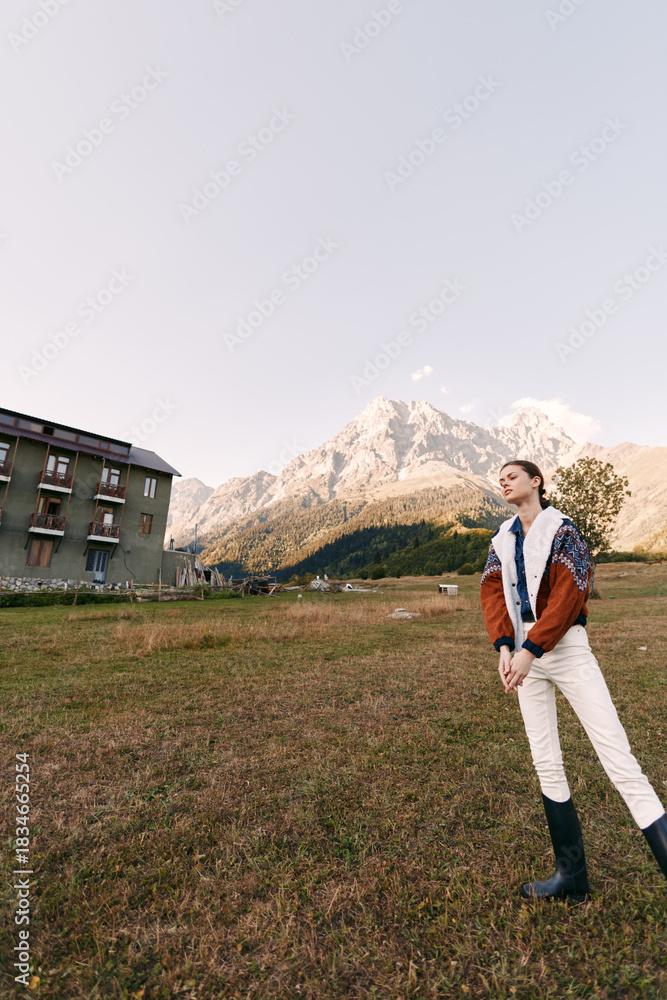 Obraz premium Woman in a jacket and boots standing in a meadow near a rustic building with alpine mountain backdrop, outdoor portrait capturing scenic landscape, sky and peaceful countryside view.